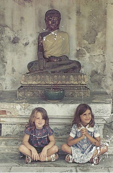 Two young, smiling girls sitting on the ground in front of a buddha statue