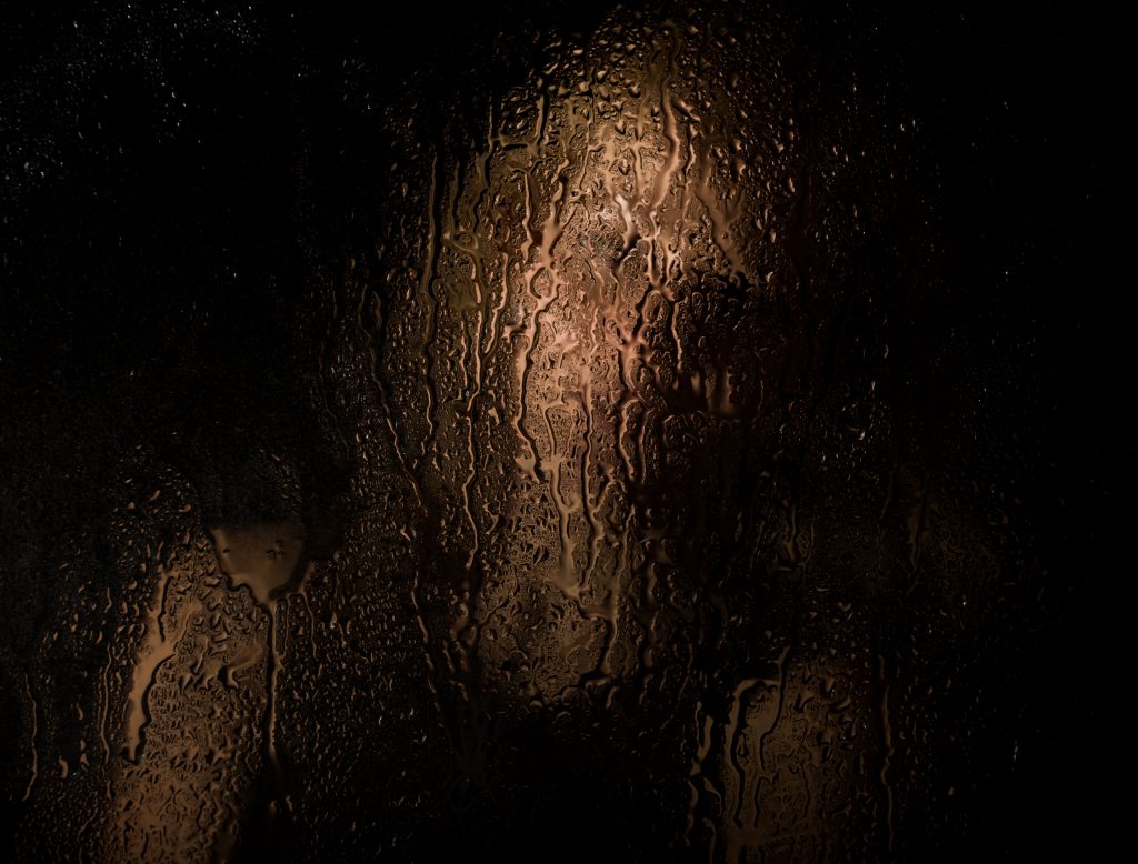 Smooth portrait of sexy model, posing behind transparent glass covered by water drops. young melancholy and sad woman portrait.